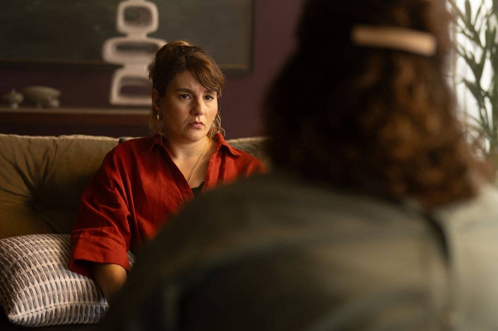 Two women sit on a couch, one in a red shirt, engaged in conversation.