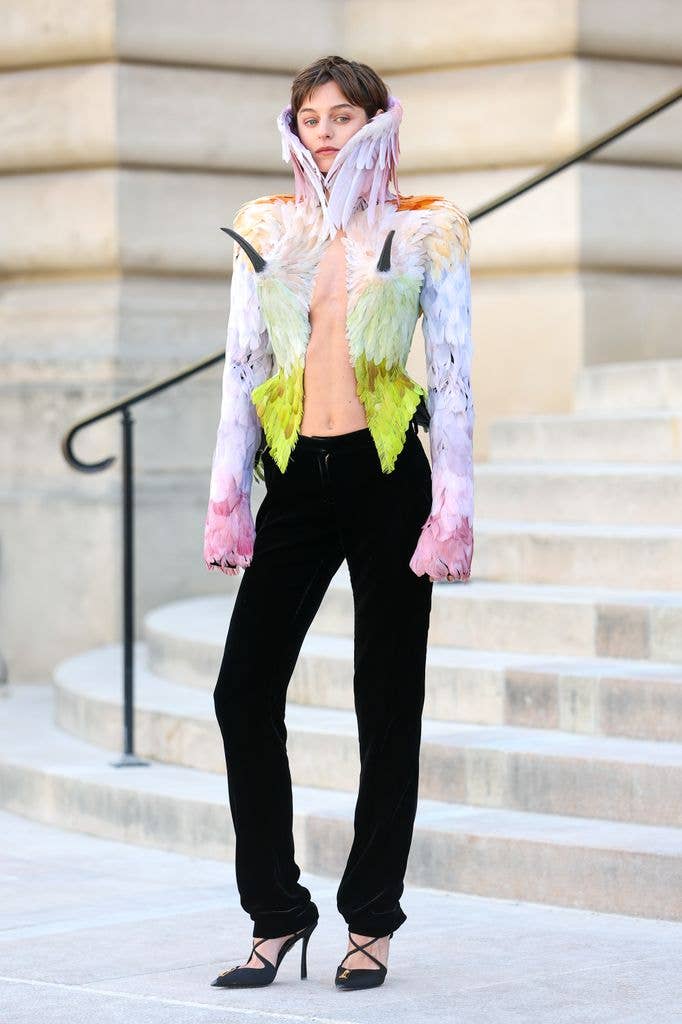Emma Corrin wears a multi coloured feathered jacket and black trousers