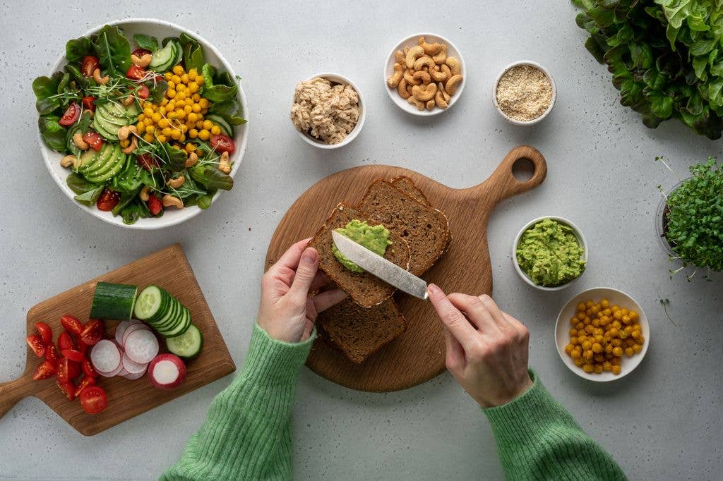 Person in green sweater slicing bread with avocado on a wooden board, surrounded by various healthy ingredients.