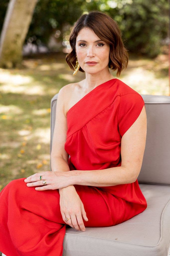 Gemma Arterton sits on a gray couch in a red dress, wearing gold earrings and rings.