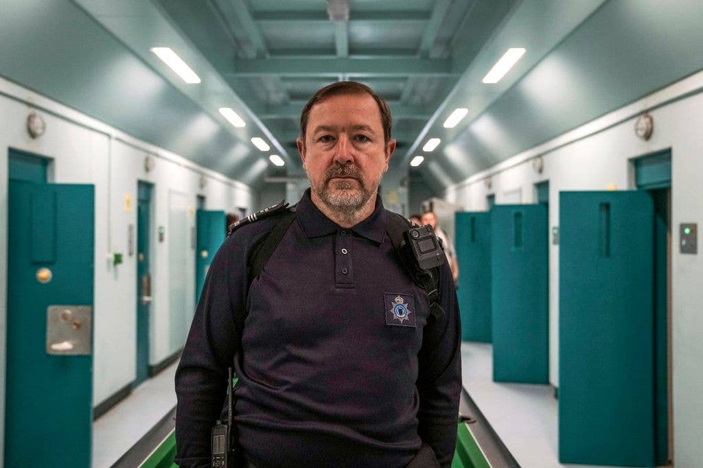 Daniel Ryan stands in a corridor with green doors, wearing a black uniform with a badge.