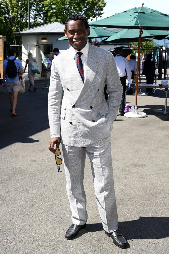 David Harewood in a light gray double-breasted suit, red and blue tie, holding sunglasses.
