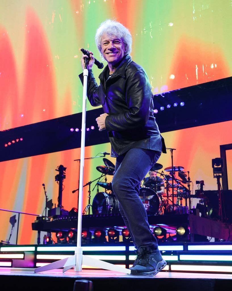 Jon Bon Jovi with white hair sings on stage with colorful backdrop and band.