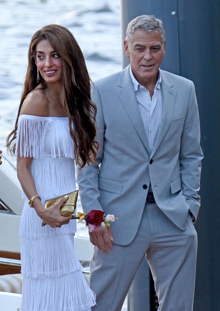 Amal wearing a white Giambattista Valli off-the-shoulder dress with fringe detailing, accessorized with a gold clutch and bracelet.