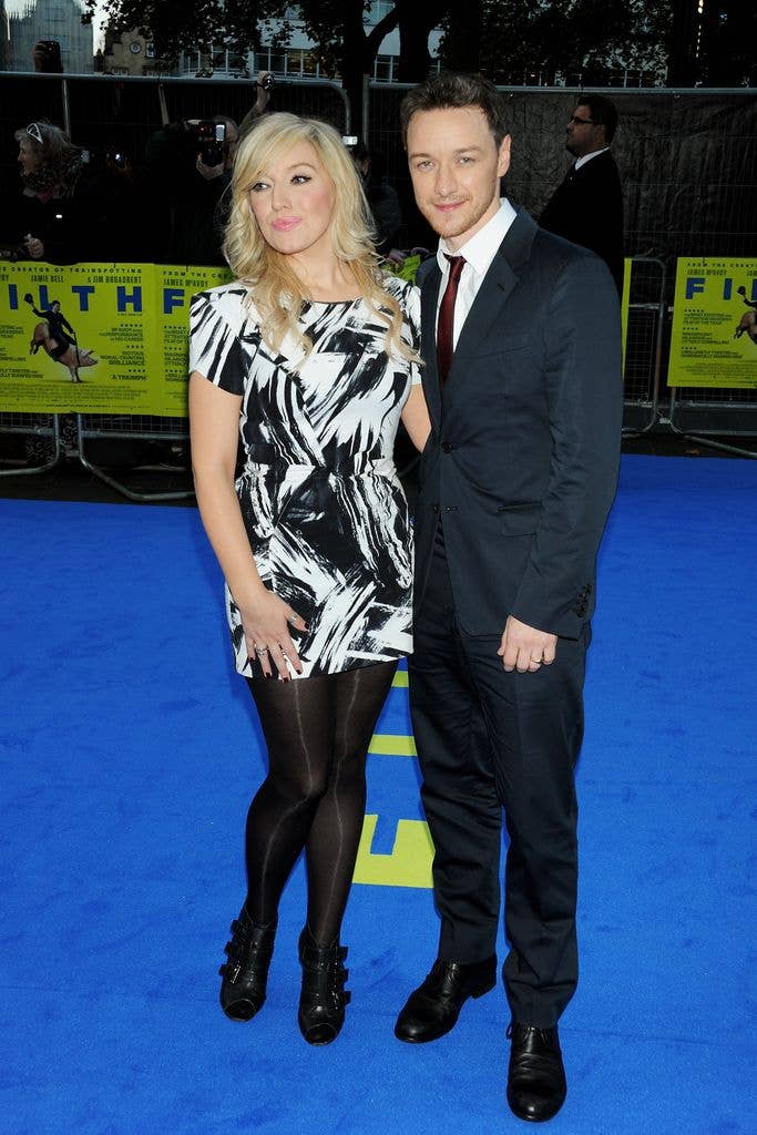 James McAvoy in a black suit and red tie stands next to a woman in a black and white dress on a blue carpet.