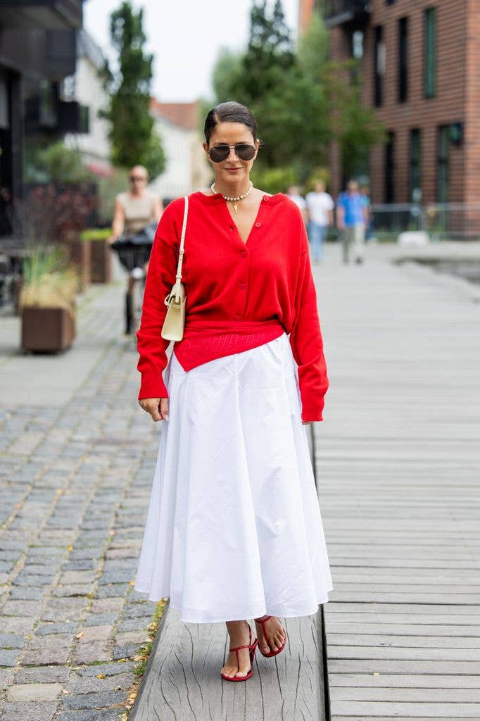 Gili Biegun wears red cardigan, white high waisted skirt, beige bag, red sandals, golden necklace, Rayban sunglasses at Copenhagen Fashion Week