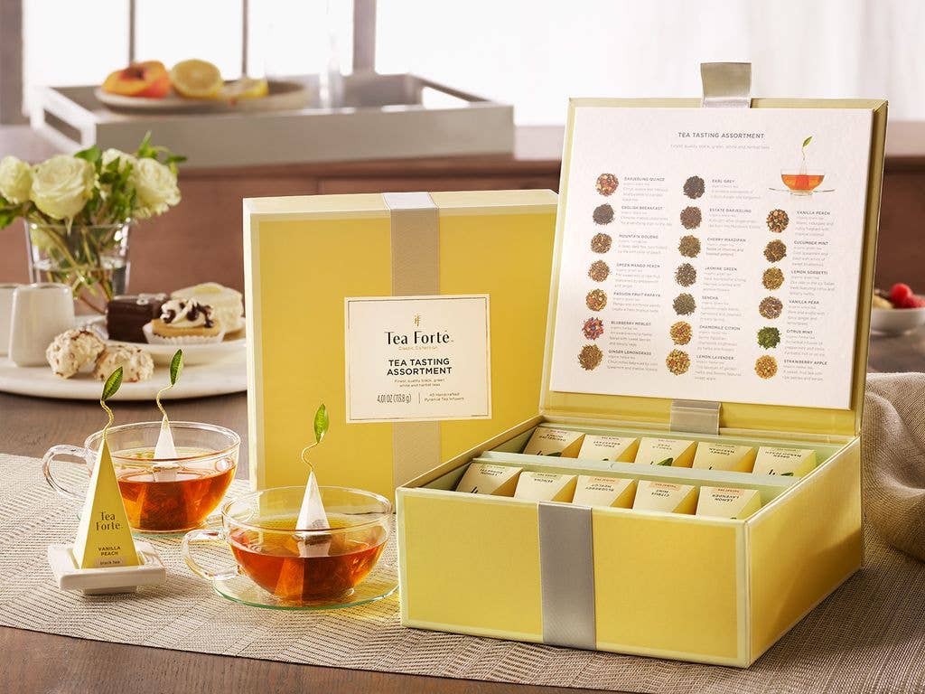 Classic Tea Tasting Assortment tea chests open and closed, teacups and pyramid infuser with tea tray, desserts and flowers in the back
