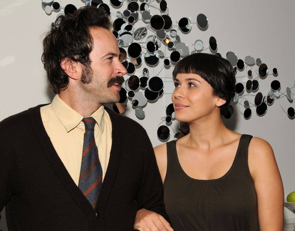 Jason Lee and a woman stand close, smiling, in front of a mirrored art piece.