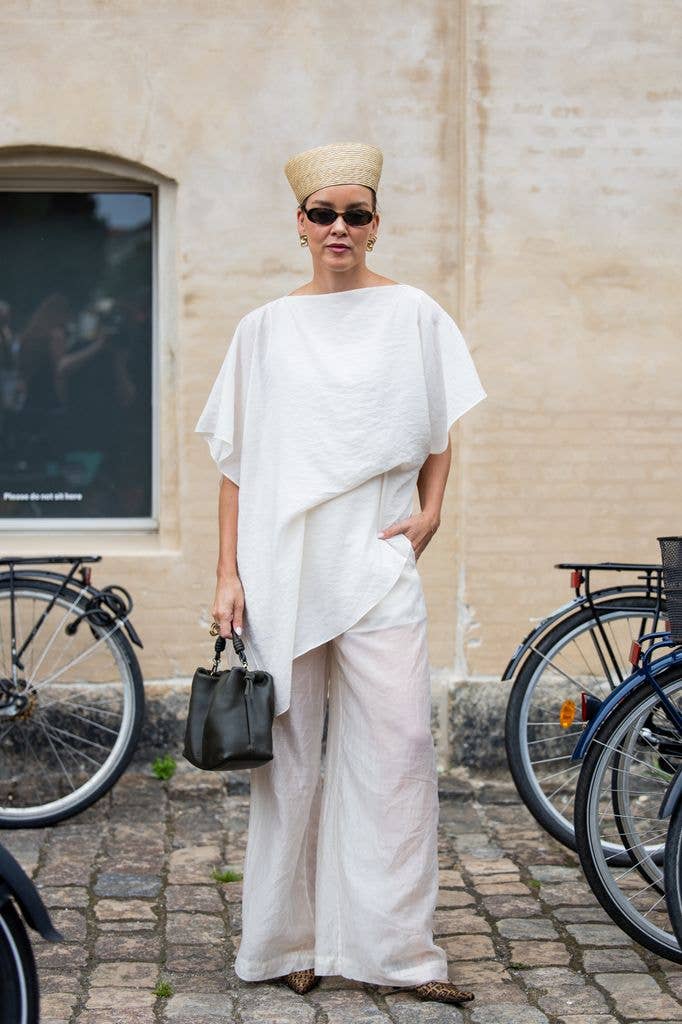 Tina Haase wears beige straw hat, white shirt, wide leg pants, black bag, sunglasses during Copenhagen Fashion Week