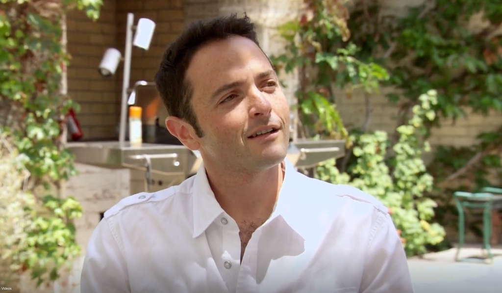 A man in a white shirt stands in a garden, smiling at the camera.