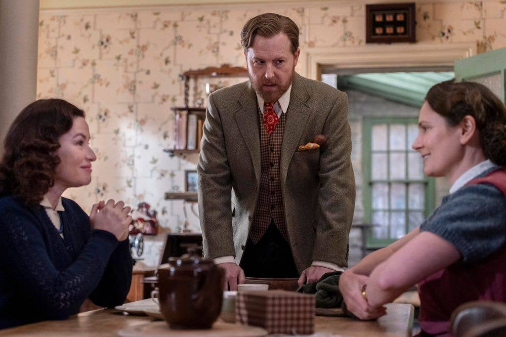 Samuel West stands at a table with two women, all engaged in conversation.