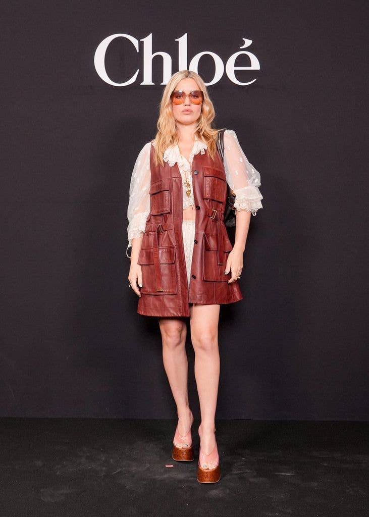Georgia May Jagger stands in front of a Chloé backdrop, wearing a red leather vest and white blouse.