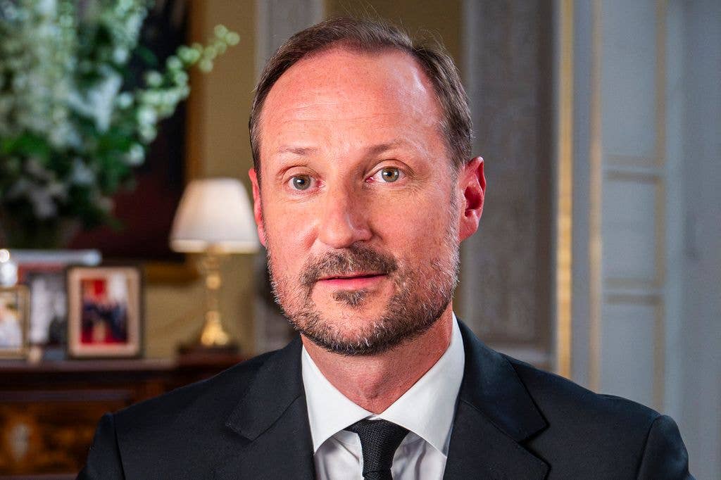 King Haakon in a black suit and white shirt, stands in an elegant room.