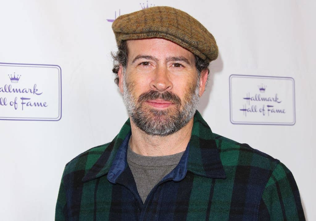 Jason Lee stands in front of a Hall of Fame backdrop, wearing a plaid shirt and flat cap.