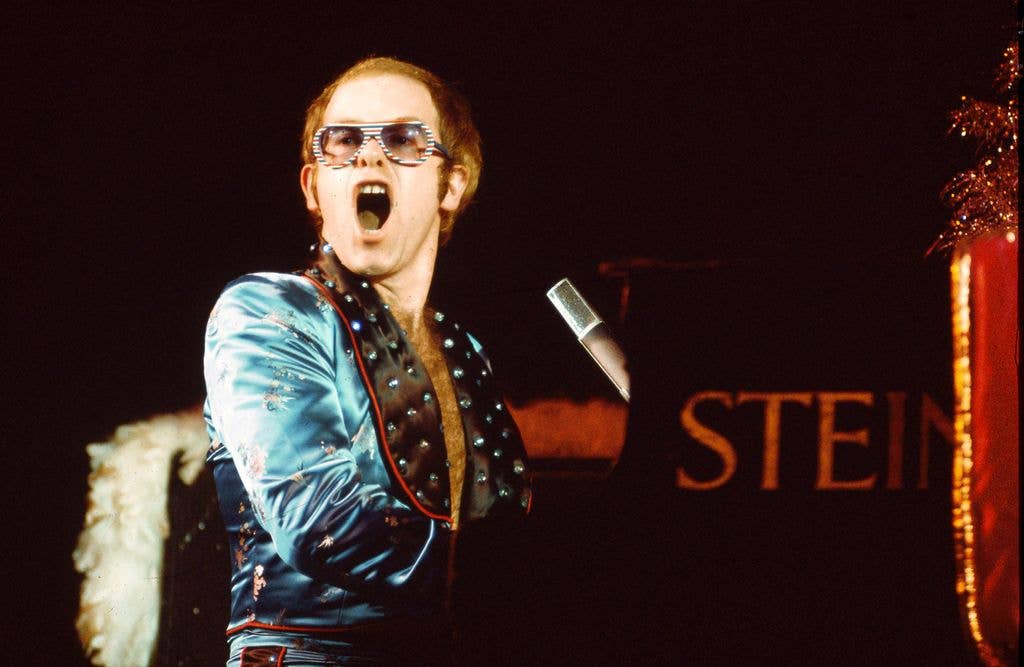 Elton John at piano in teal robe and colourful sunglasses.