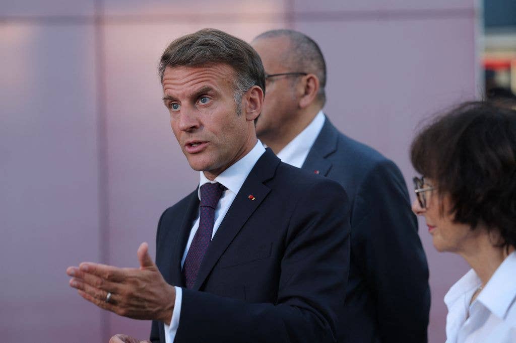Emmanuel Macron, in a dark suit and red tie, gestures while speaking to a woman in a white shirt.