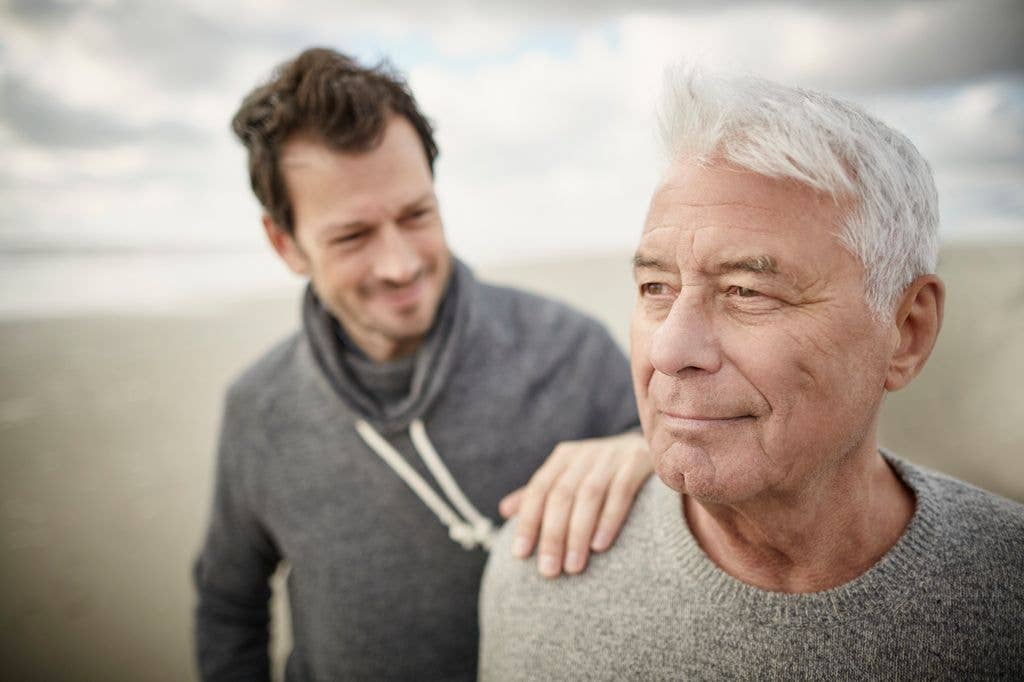 Two men on a beach, one with white hair in a gray sweater, the other in a gray hoodie.