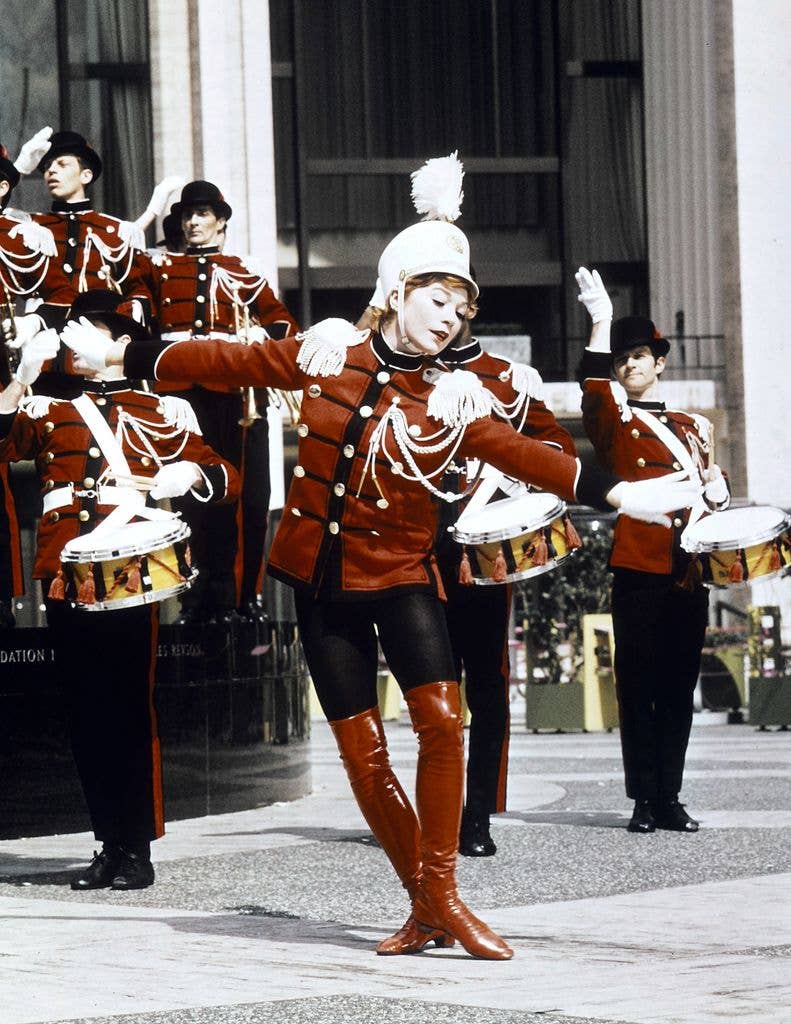 Shirley MacLaine in a red uniform and white hat, dancing with a drum, surrounded by band members.
