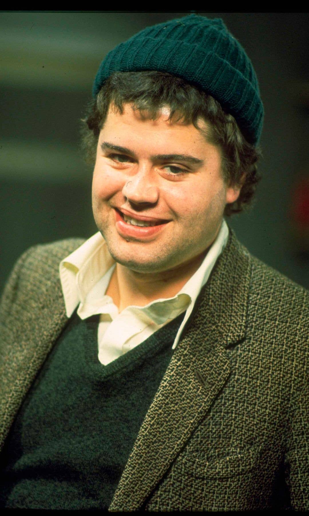 Paul Henry as Benny, smiling, wearing a green beanie and a gray jacket.