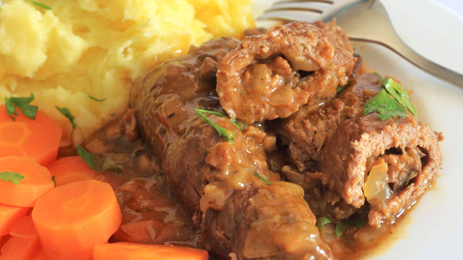 Plate with beef rouladen, mashed potatoes, and sliced carrots with a fork.