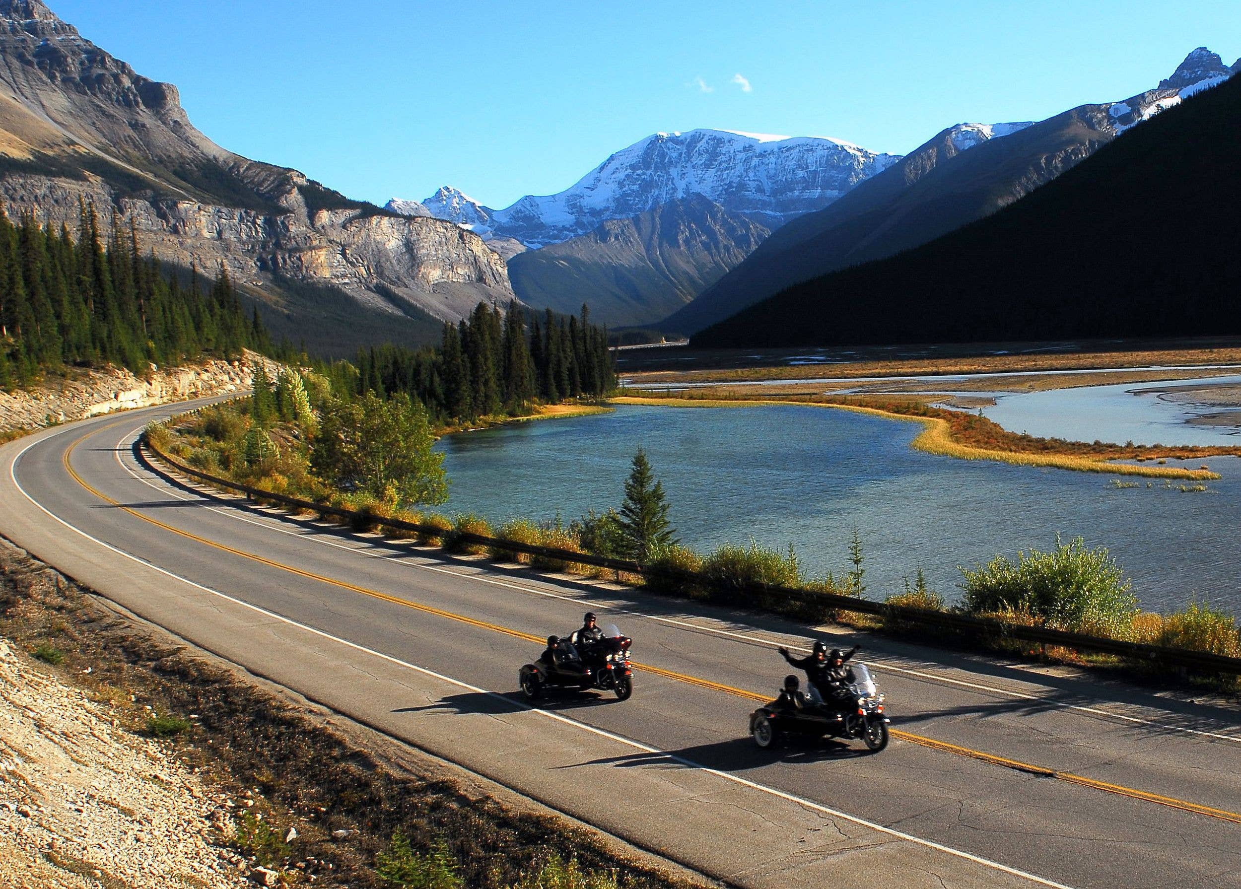 Two motorcycles drive along the road in Jasper National Park with mountains and a river in the backdrop