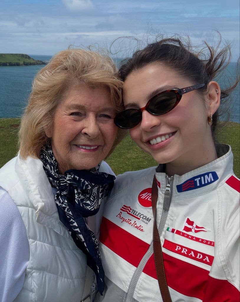 Two smiling women stand by the ocean, one in a white vest and scarf, the other in a racing suit with 'TIM' and 'PRADA' logos.