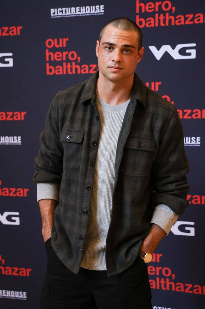 Noah Centineo stands with hands in pockets, wearing a plaid jacket and gray sweater.