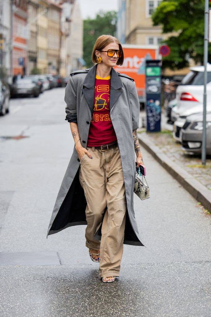 Marianne Theodorsen wears earrings, sunglasses, oversized grey trench coat, beige pants, red yellow shirt with print outside MKDT Studio during the Copenhagen Fashion Week