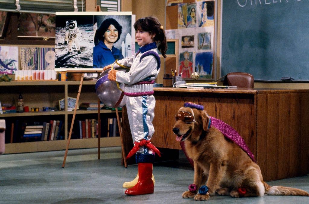 Soleil Moon Frye as Penelope Brewster giving a presentation dressed as an astronaut, stood next to her dog at the front of the classroom.