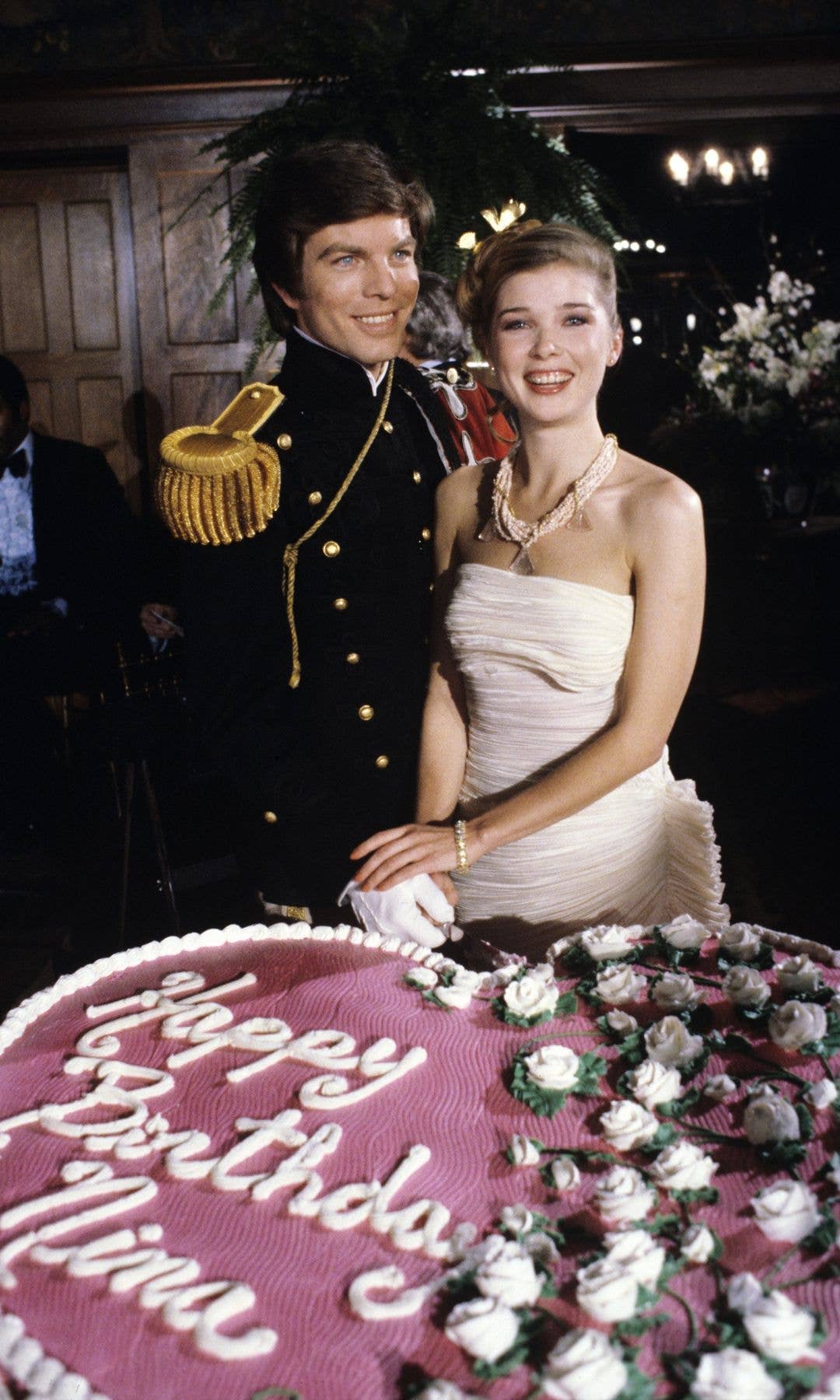 Man and woman in formal attire stand by a birthday cake, smiling.