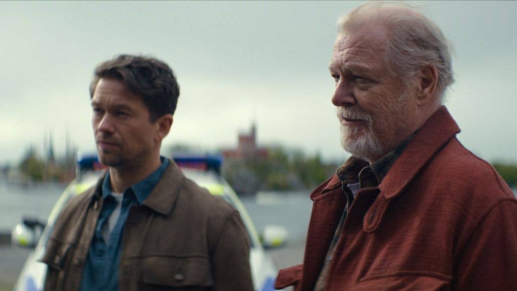 Jakob Oftebro stands next to an older man with a white beard, both looking ahead. A police car is behind them.
