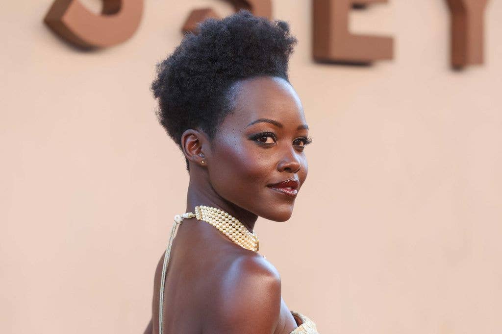 Lupita Nyong'o in a strapless dress with a pearl necklace, standing against a beige background.