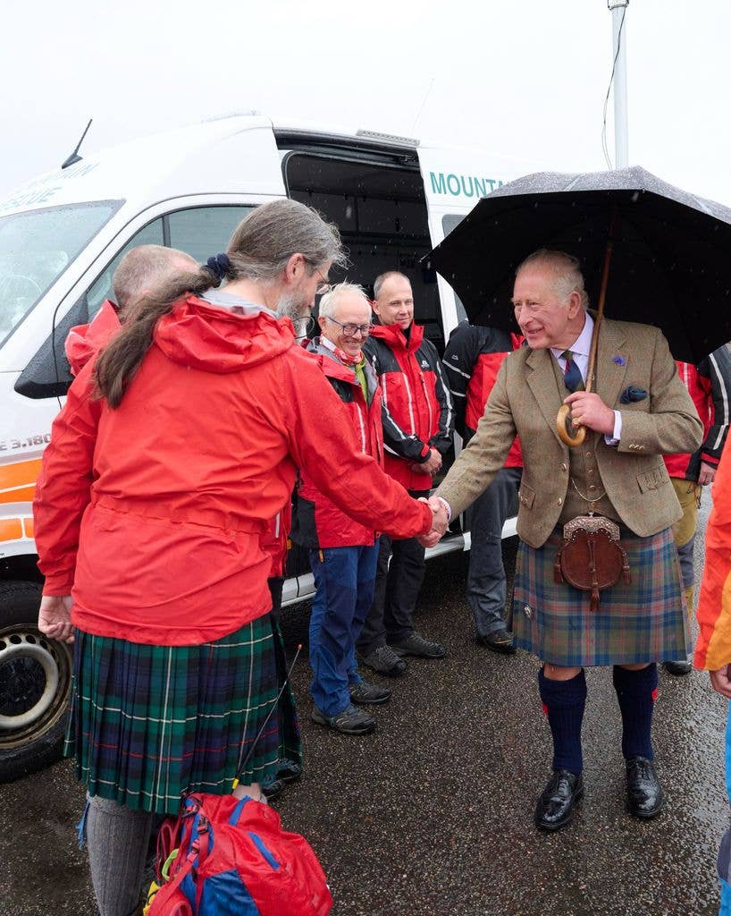King Charles in a kilt and holding an umbrella, shakes hands with a woman in a red jacket.