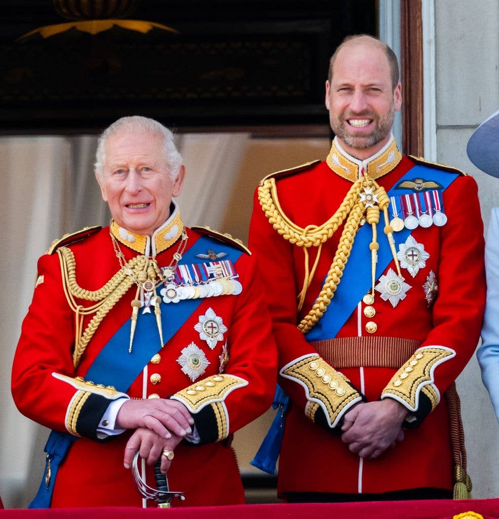 Prince Charles and Prince William stand together in red military uniforms with medals.