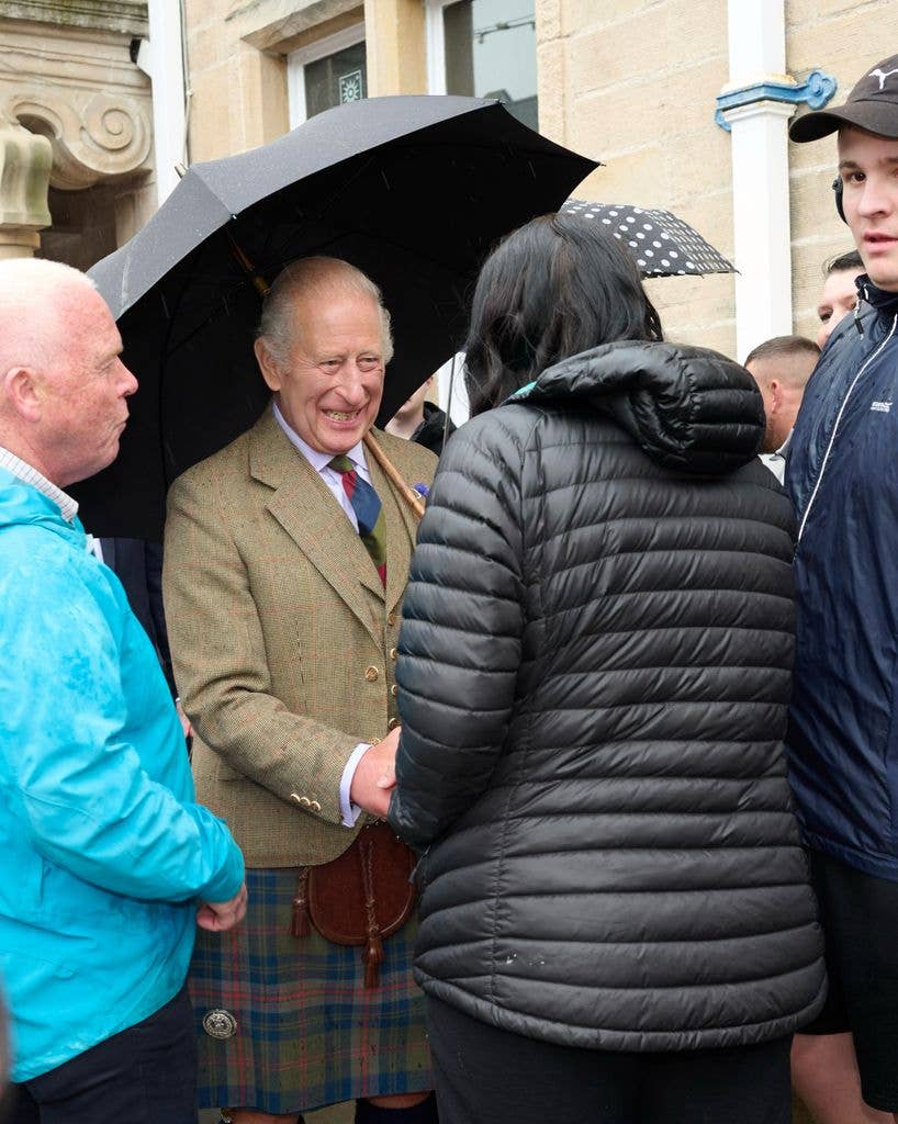 King Charles in a plaid kilt and holding an umbrella, shakes hands with a woman in a black jacket.