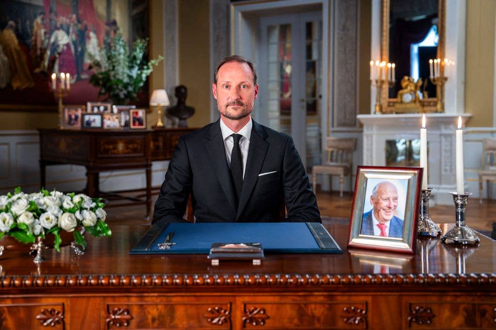 King Haakon in formal attire at a desk with a portrait of Harald V of Norway.