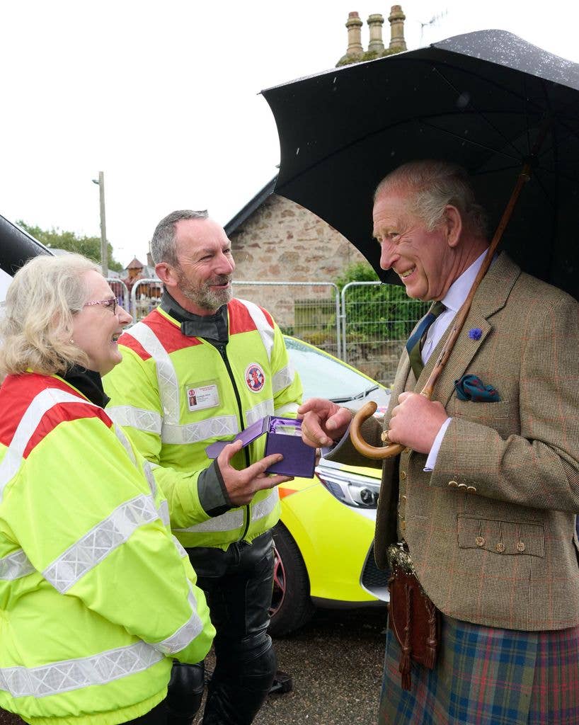 King Charles holding an umbrella and a gift box, stands with two individuals in high-visibility jackets.