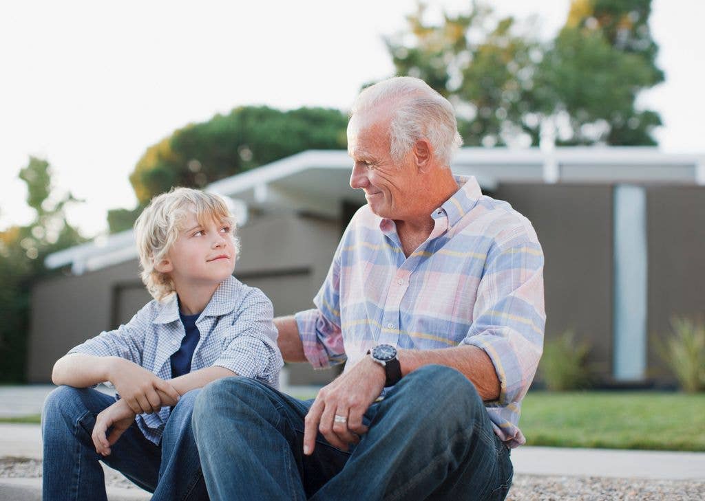 An older man with a watch and a young boy sit on a curb, smiling.