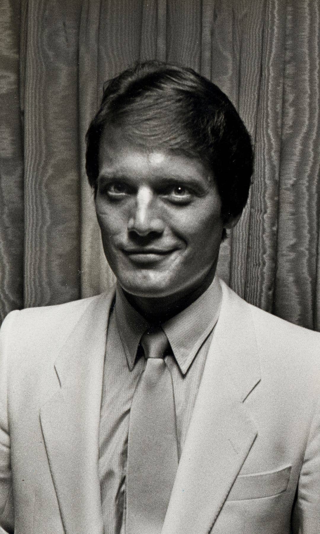 A smiling man in a white suit and tie stands in front of a curtain.