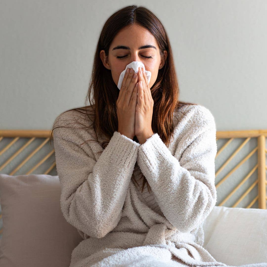 Young caucasian woman blowing nose with paper tissue wearing a robe sitting on bed suffering from a cold. Cold and sickness concept.