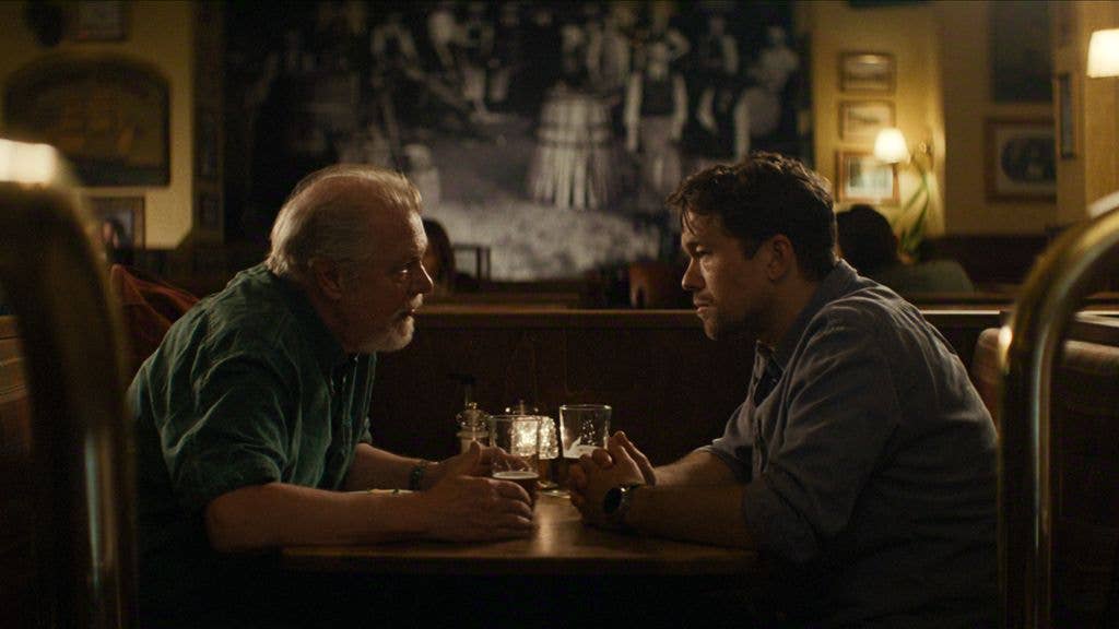Two men, one with short hair and the other with gray hair, sit at a table in a restaurant, facing each other.
