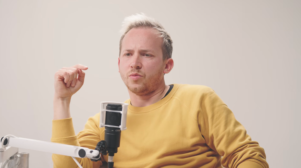 Mike Bailey in a yellow sweater speaks into a microphone, hand raised.