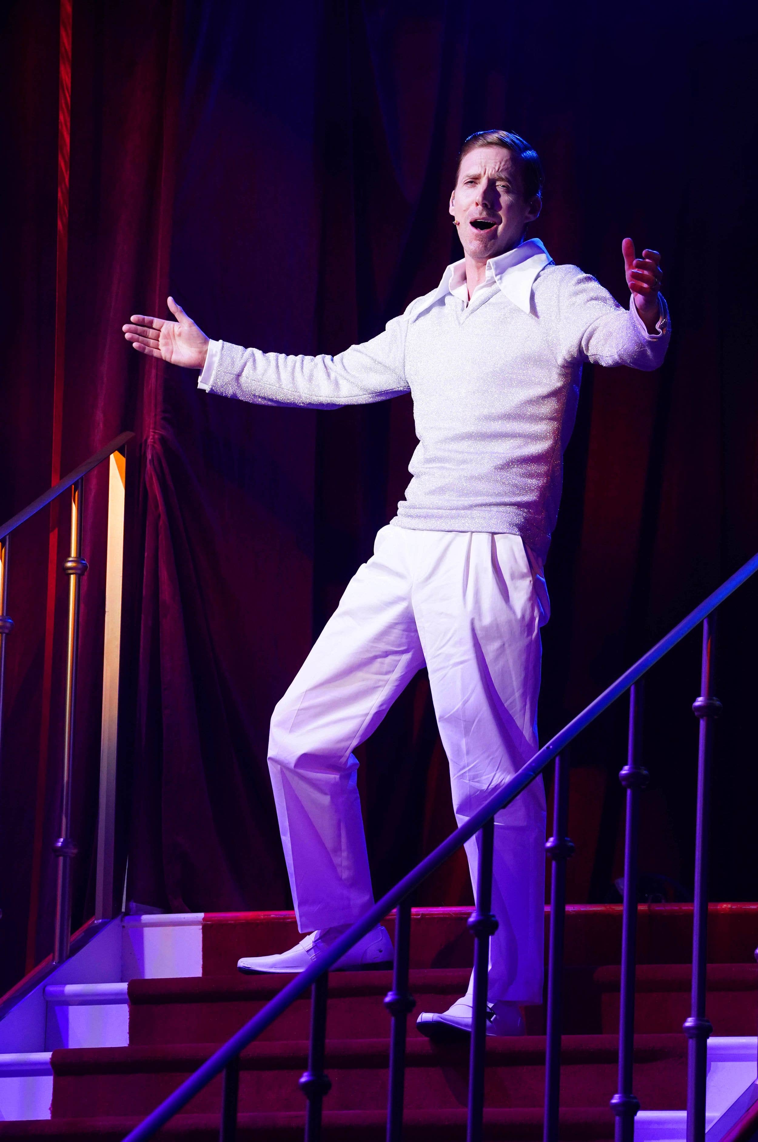Ricky Wilson, dressed in white and standing on a staircase with arms wide, plays Teen Angel in Grease: The Immersive Movie Musical, at Evolution London in July 2026