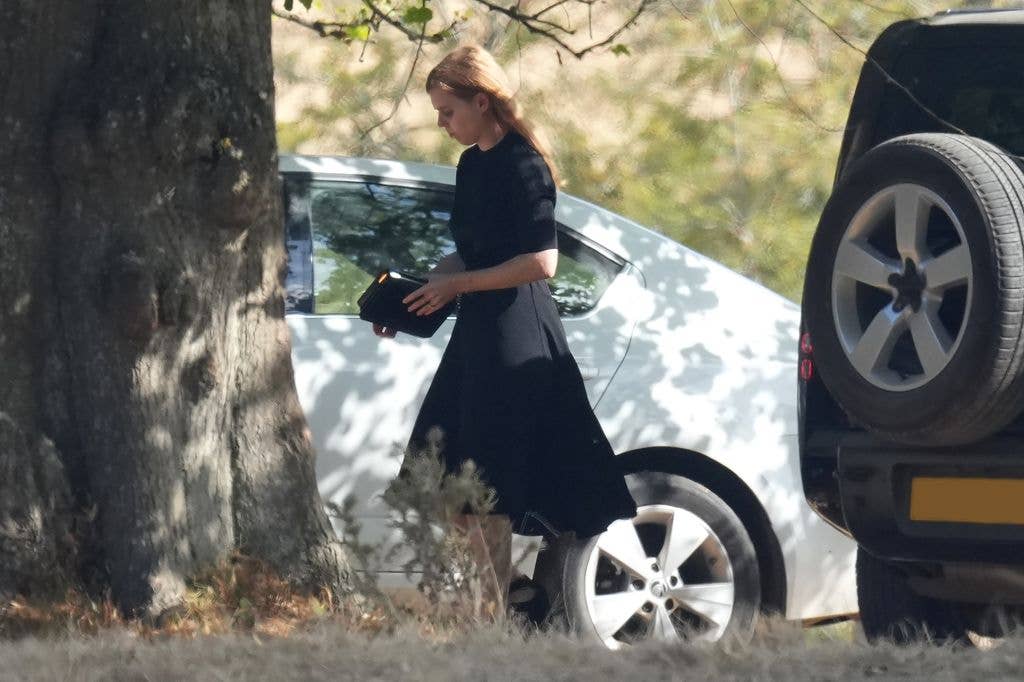 Princess Beatrice in a black dress walks by a white car, holding a black bag.