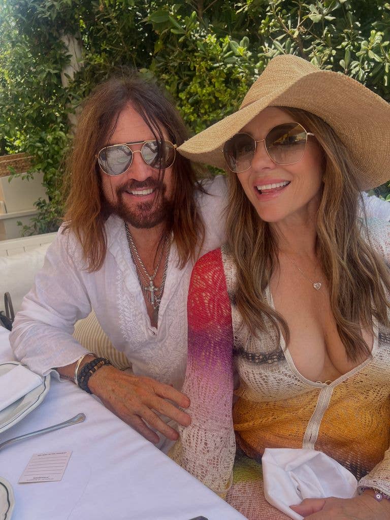 Billy Ray Cyrus and Elizabeth Hurley sitting together, smiling, wearing sunglasses.