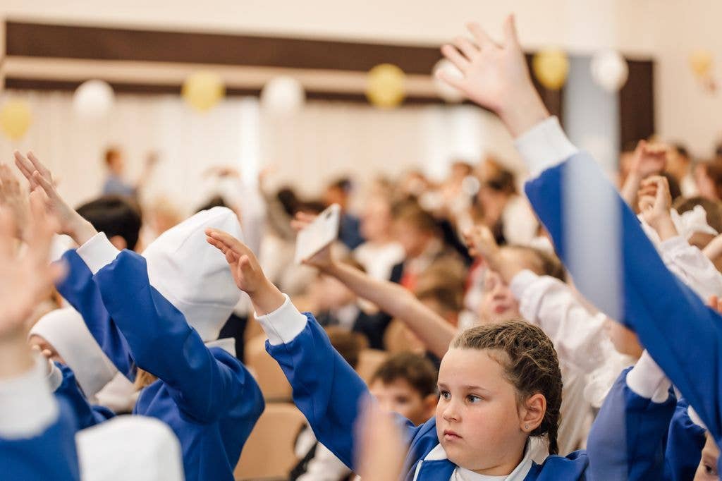 Children in blue uniforms and white hats raise their hands in a large hall with balloons.