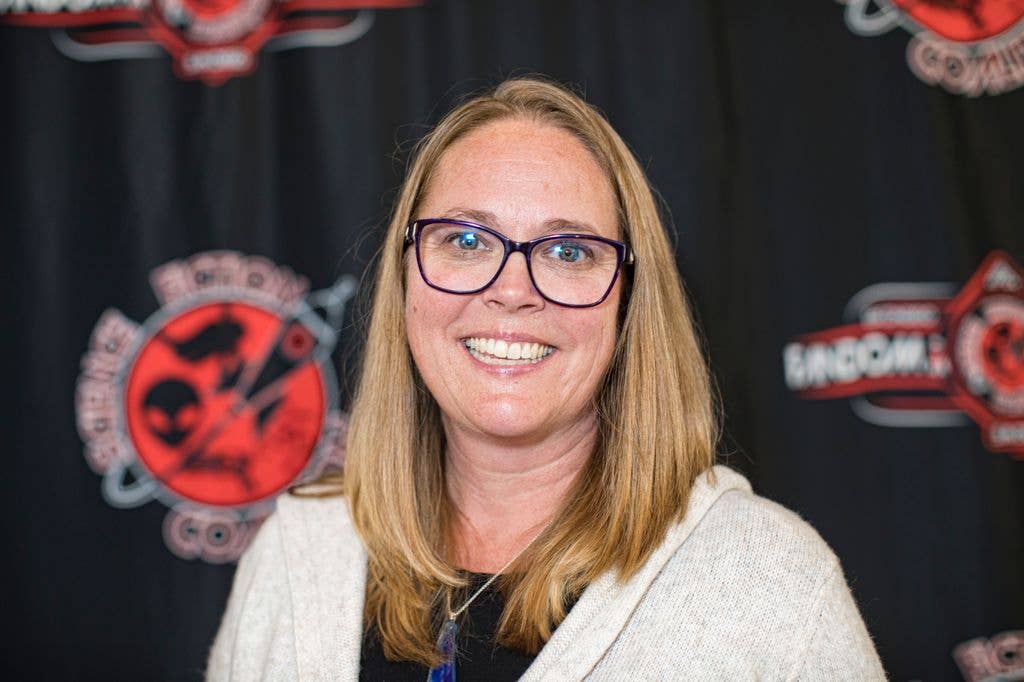A smiling woman with blonde hair wearing glasses and a white cardigan stands in front of a black backdrop.
