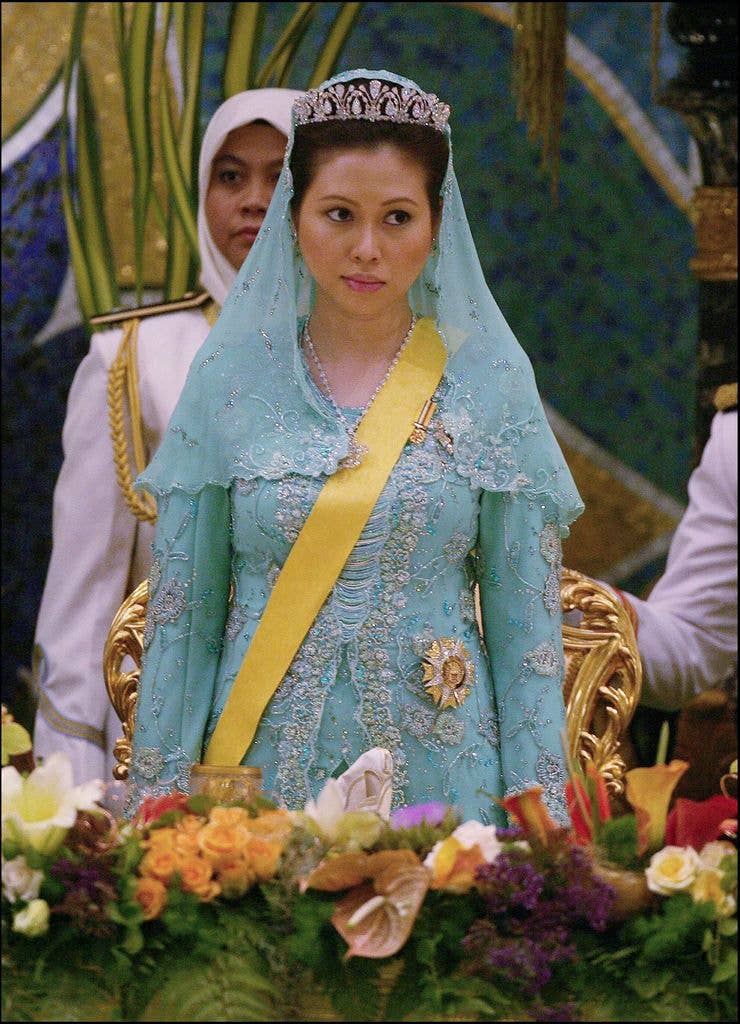 A woman in a blue gown and crown stands in front of a floral arrangement.