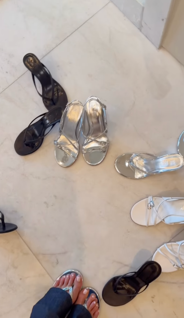 Person wearing silver sandals stands among various sandals on a tiled floor.