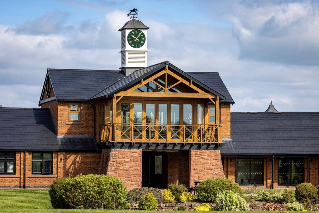 Large brick building with a clock tower and balcony, set in a green landscape with bushes.